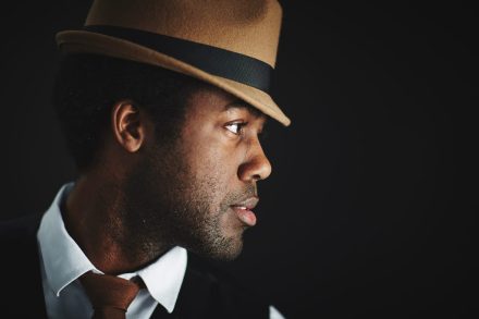 A man in profile wearing a tan fedora with a black band, looking to the right. He has a thoughtful expression, dressed in a white shirt, vest, and brown tie. The background is dark, highlighting his face and hat.
