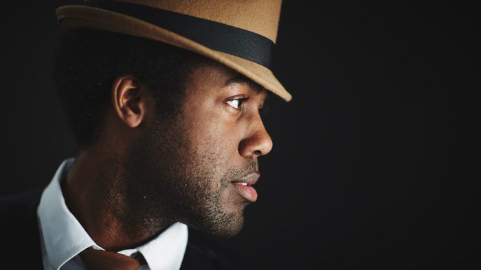 A man in profile wearing a tan fedora with a black band, looking to the right. He has a thoughtful expression, dressed in a white shirt, vest, and brown tie. The background is dark, highlighting his face and hat.