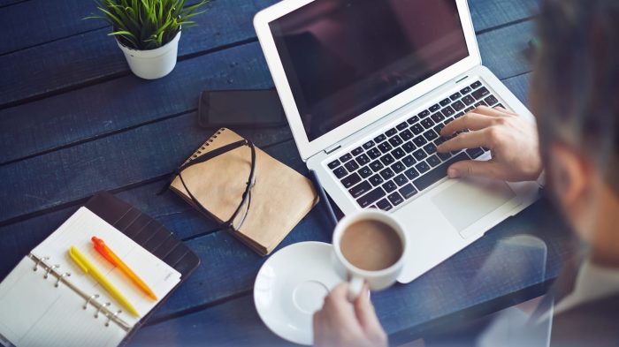 A person works on a laptop at a blue wooden table, holding a cup of coffee. Nearby are an open notebook with pens, a closed journal, a smartphone, and a small plant. The scene suggests a workspace or study area.