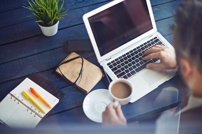 A person works on a laptop at a blue wooden table, holding a cup of coffee. Nearby are an open notebook with pens, a closed journal, a smartphone, and a small plant. The scene suggests a workspace or study area.