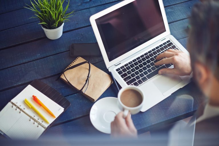 A person works on a laptop at a blue wooden table, holding a cup of coffee. Nearby are an open notebook with pens, a closed journal, a smartphone, and a small plant. The scene suggests a workspace or study area.