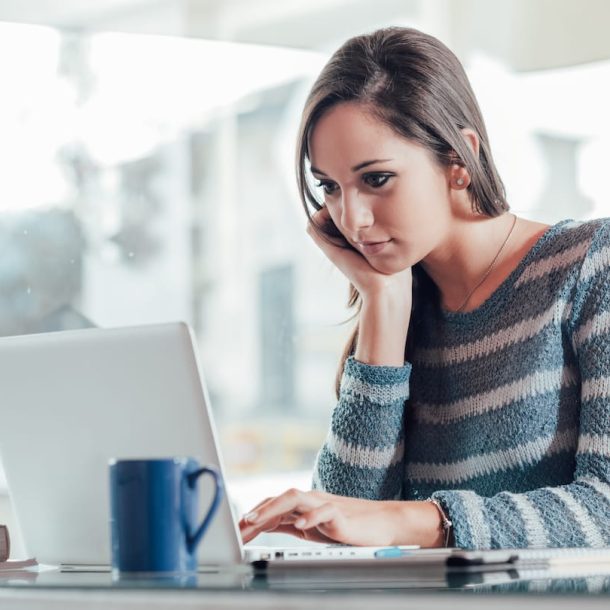 A woman in a striped sweater sits at a table, looking at a laptop. She rests her chin on her hand and holds a phone to her ear. A blue mug and notebooks are on the table. The background shows a bright interior space.