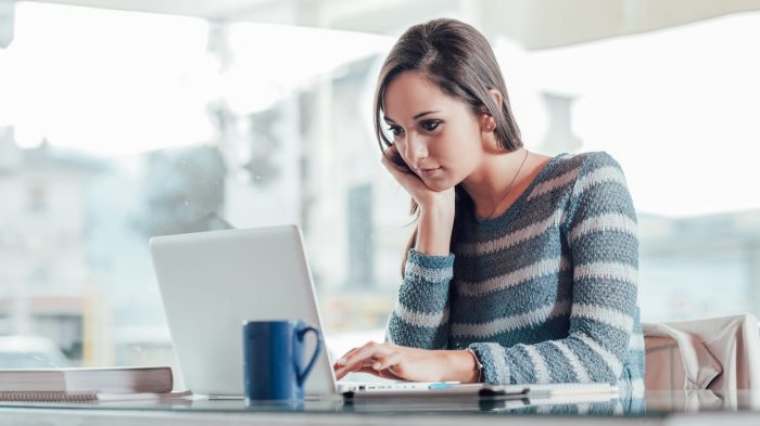 A woman in a striped sweater sits at a table, looking at a laptop. She rests her chin on her hand and holds a phone to her ear. A blue mug and notebooks are on the table. The background shows a bright interior space.