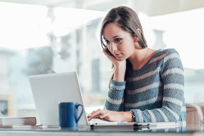 A woman in a striped sweater sits at a table, looking at a laptop. She rests her chin on her hand and holds a phone to her ear. A blue mug and notebooks are on the table. The background shows a bright interior space.