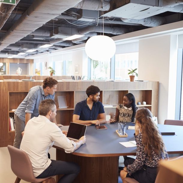 Five people are gathered around a round table in a modern, open office. They are engaged in a discussion, with laptops and documents on the table. Shelves with books and plants are in the background, and large windows let in natural light.