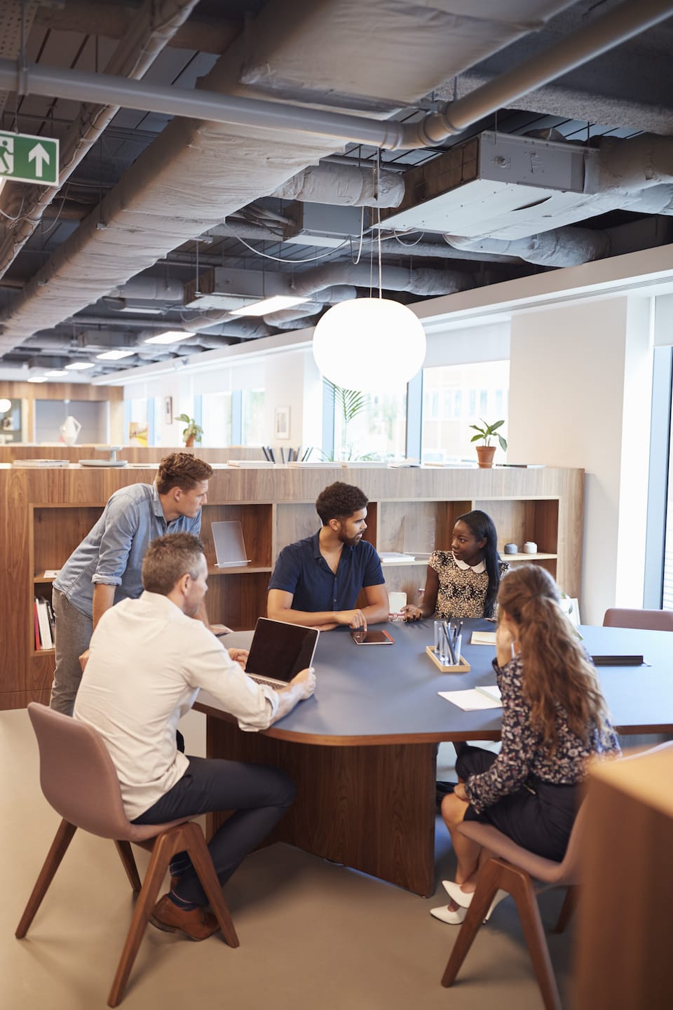 Five people are gathered around a round table in a modern, open office. They are engaged in a discussion, with laptops and documents on the table. Shelves with books and plants are in the background, and large windows let in natural light.