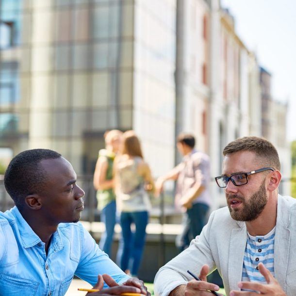 Two men sit at an outdoor table, talking over coffee with notebooks and smartphones in front of them. The man on the left wears a light blue shirt, while the man on the right wears glasses and a blazer. Blurred background of people and buildings.