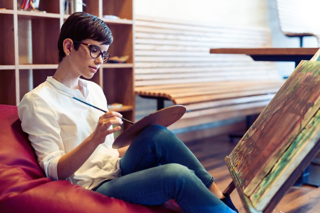 A person with short hair and glasses sits on a red beanbag, painting on a canvas propped up in front of them. They hold a paintbrush and palette, focused on their work. The background includes wooden shelves and a table.