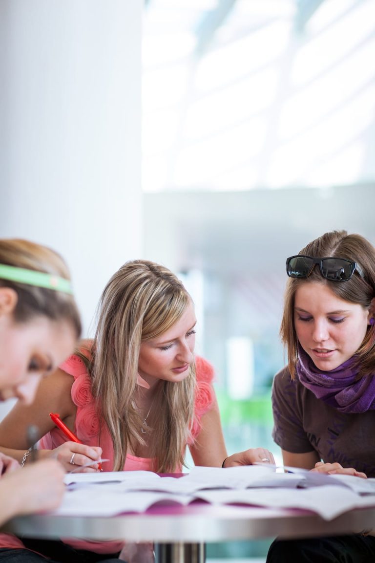 Three people sit at a table, focused on studying papers spread out before them. The central figure with blonde hair leans in to discuss, while another with sunglasses on her head looks at the papers. The background is softly blurred.