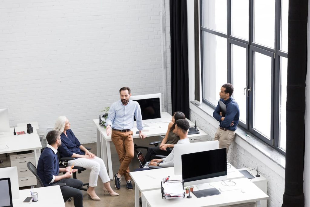 A group of six people is having a discussion in a modern office. They are gathered around a desk with computers and a laptop. Large windows allow natural light to illuminate the space. The atmosphere is collaborative and professional.