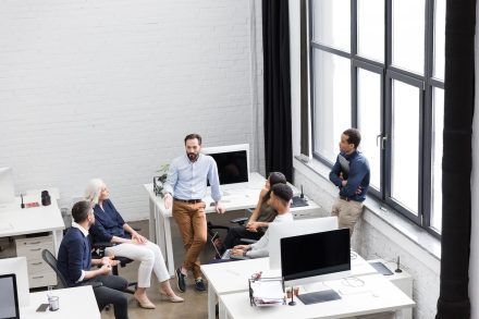 A group of six people is having a discussion in a modern office. They are gathered around a desk with computers and a laptop. Large windows allow natural light to illuminate the space. The atmosphere is collaborative and professional.