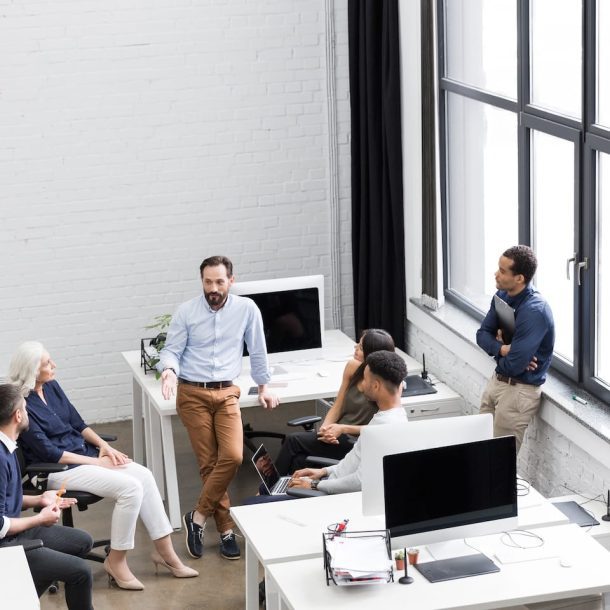 A group of six people is having a discussion in a modern office. They are gathered around a desk with computers and a laptop. Large windows allow natural light to illuminate the space. The atmosphere is collaborative and professional.