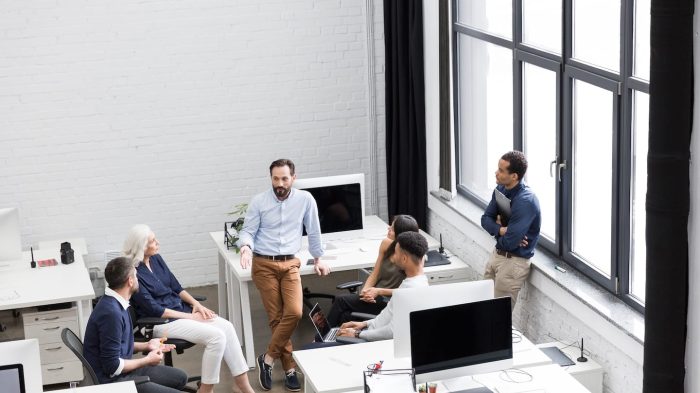 A group of six people is having a discussion in a modern office. They are gathered around a desk with computers and a laptop. Large windows allow natural light to illuminate the space. The atmosphere is collaborative and professional.