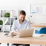 A man sits at a desk working on a laptop in a bright office. Papers, a smartphone, and a coffee cup are on the desk. Behind him is a shelf with binders and decor. The setting is modern and tidy.