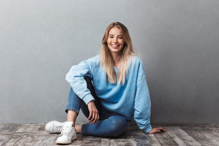 Smiling person with long blonde hair sitting on a wooden floor against a gray wall. They wear a light blue sweater, rolled-up jeans, and white sneakers, with one hand on the floor and the other resting on their knee.