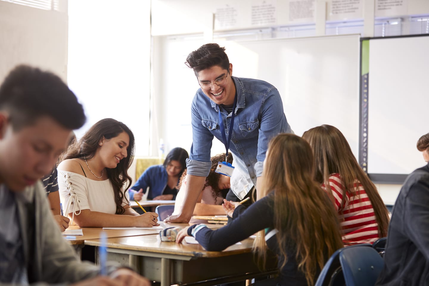 A teacher leans over a desk, engaging with a group of students in a classroom. The students are seated and writing at their desks, while the teacher smiles and interacts with them. The classroom has bright lighting and educational posters.