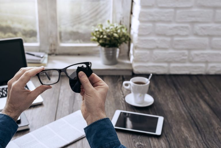 A person cleans eyeglasses with a cloth at a wooden desk. Nearby are a laptop, a newspaper, a tablet, and a cup of coffee. A small potted plant is on the windowsill, with natural light coming through the window.