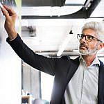 A person with gray hair and glasses is writing on a whiteboard in an office setting. They are wearing a suit jacket over a light-colored shirt. The room has modern decor with exposed ceilings and bright lighting.