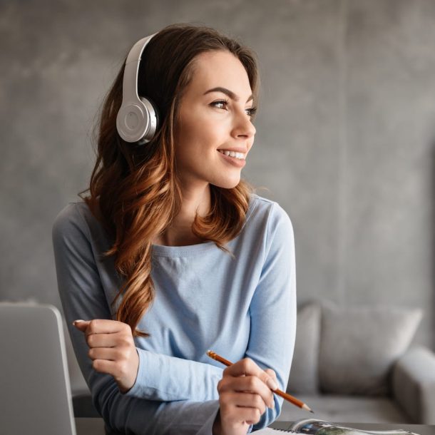 A woman wearing headphones looks to the side while sitting at a desk with a laptop and open books. She holds a pencil and wears a light blue sweater, appearing thoughtful and engaged. The background is a gray wall and a couch.