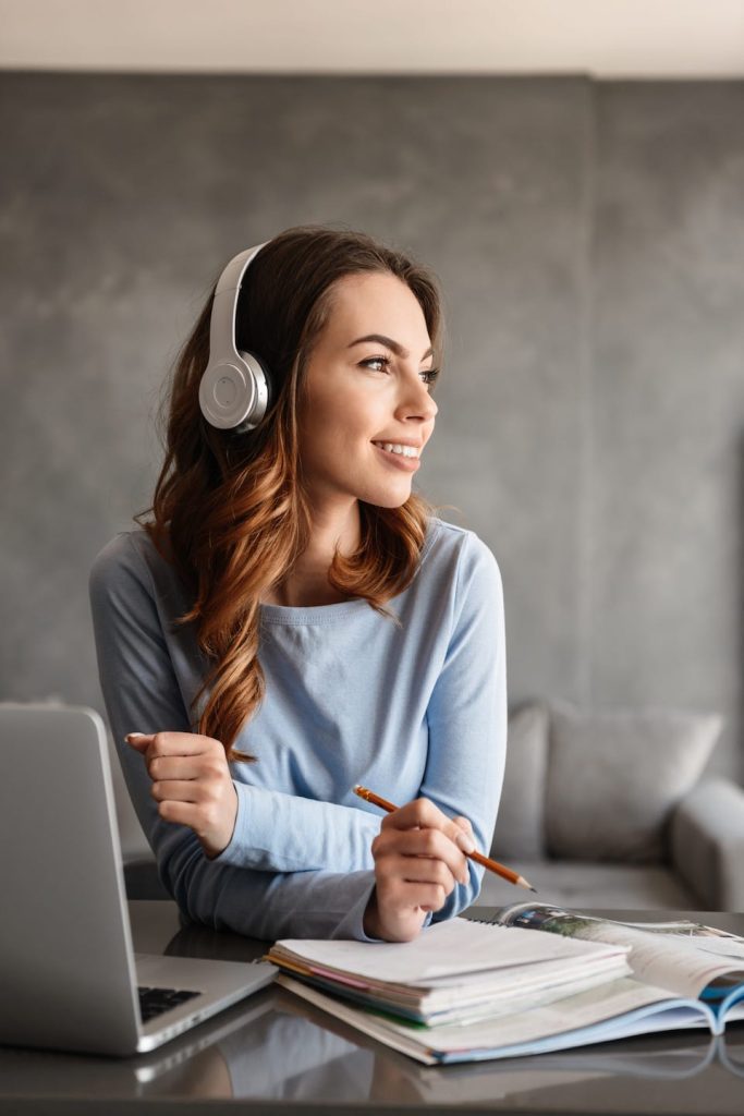 A woman wearing headphones looks to the side while sitting at a desk with a laptop and open books. She holds a pencil and wears a light blue sweater, appearing thoughtful and engaged. The background is a gray wall and a couch.