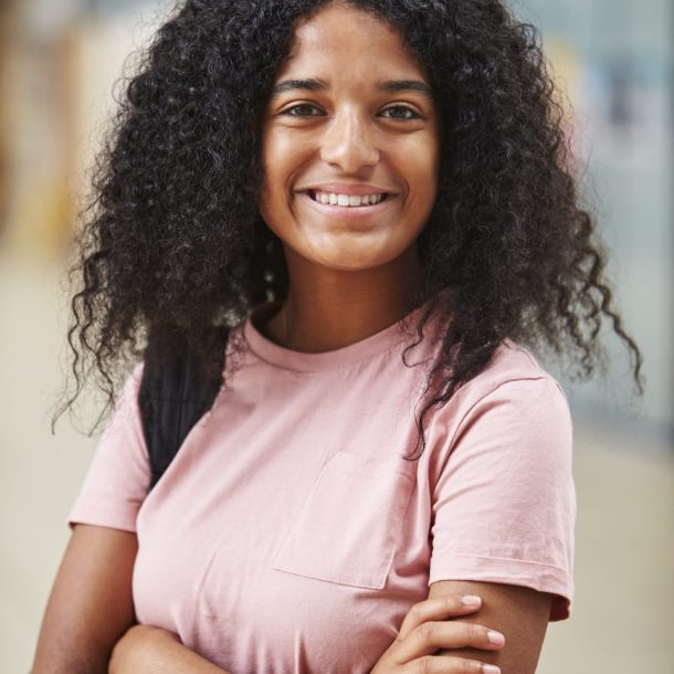 A person with curly hair and a pink t-shirt smiles confidently, arms crossed, perhaps contemplating their goals. They stand in an indoor setting, where the background details blur into insignificance.