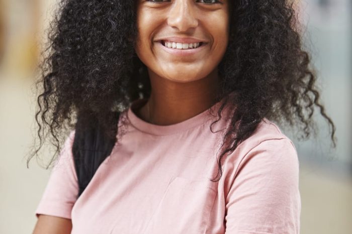 A person with curly hair and a pink t-shirt smiles confidently, arms crossed, perhaps contemplating their goals. They stand in an indoor setting, where the background details blur into insignificance.