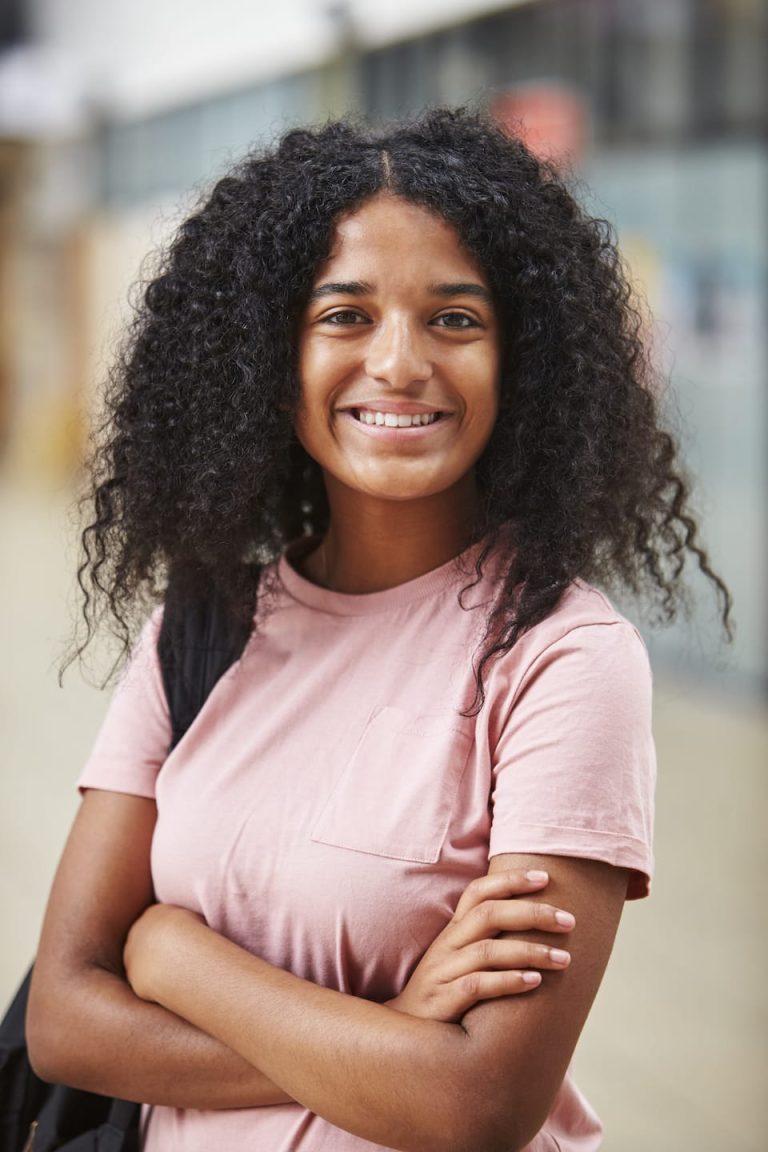A person with curly hair and a pink t-shirt smiles confidently, arms crossed, perhaps contemplating their goals. They stand in an indoor setting, where the background details blur into insignificance.