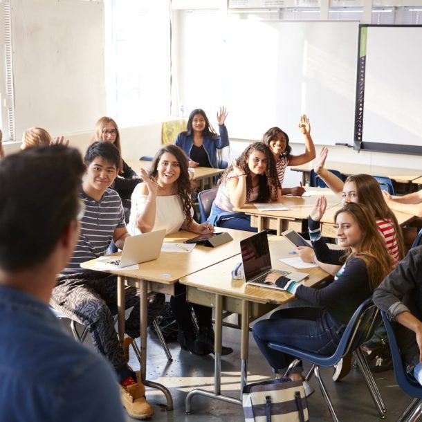 A diverse group of students in a classroom, sitting at tables, facing a teacher. Some students have their hands raised, and others are using laptops. The room is bright with large windows and a whiteboard at the front.