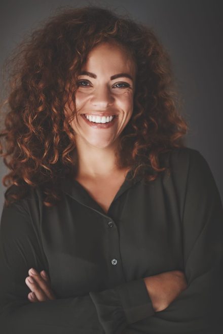 A woman with curly hair smiles warmly, wearing a dark green button-up shirt. She stands with her arms crossed against a dark background.