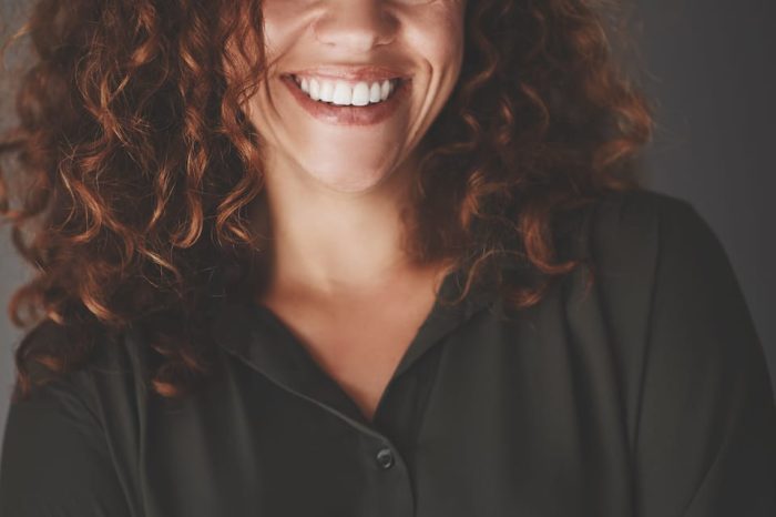 A woman with curly hair smiles warmly, wearing a dark green button-up shirt. She stands with her arms crossed against a dark background.