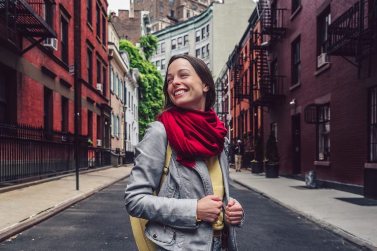 A woman with a red scarf and gray jacket smiles while strolling down a narrow urban street lined with red brick buildings. She carries a light-colored bag like shes perfectly in sync with her content calendar, appearing cheerful on the quiet, empty street.