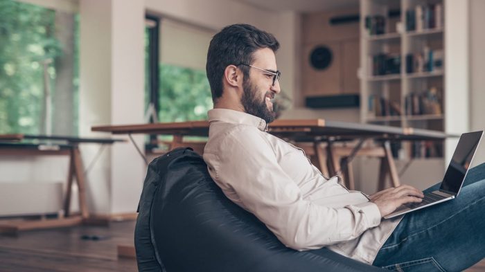 A man with a beard and glasses sits on a bean bag chair, working on a laptop. He is in a modern, well-lit room with large windows and bookshelves in the background.