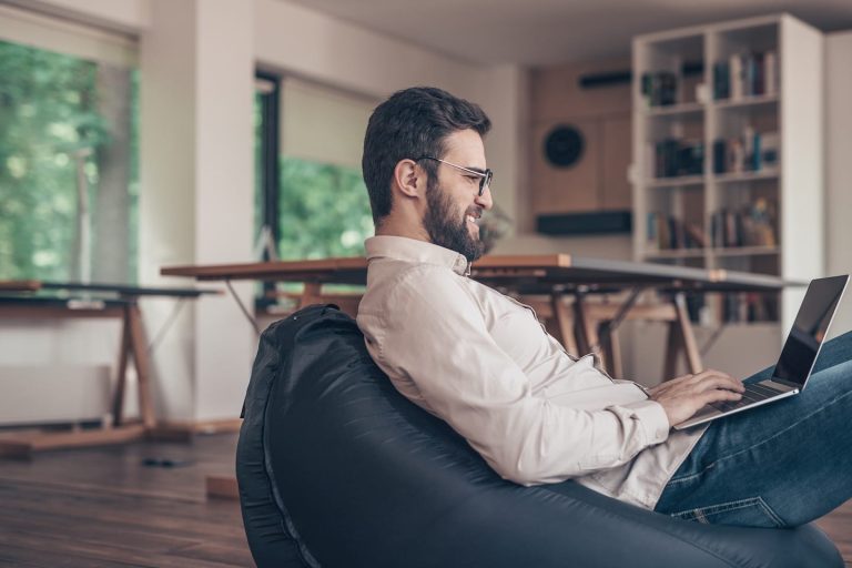A man with a beard and glasses sits on a bean bag chair, working on a laptop. He is in a modern, well-lit room with large windows and bookshelves in the background.