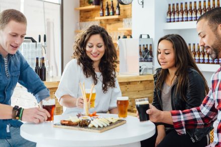 Four people sitting around a table with drinks and a platter of snacks in a casual setting. The table has various glasses of beer and a wooden board with cheese and charcuterie. Shelves with bottles are in the background.