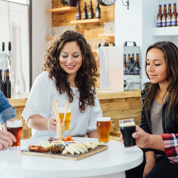 Four people sitting around a table with drinks and a platter of snacks in a casual setting. The table has various glasses of beer and a wooden board with cheese and charcuterie. Shelves with bottles are in the background.