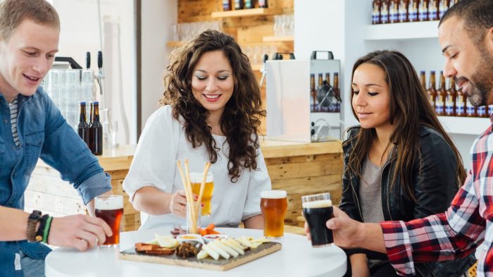 Four people sitting around a table with drinks and a platter of snacks in a casual setting. The table has various glasses of beer and a wooden board with cheese and charcuterie. Shelves with bottles are in the background.