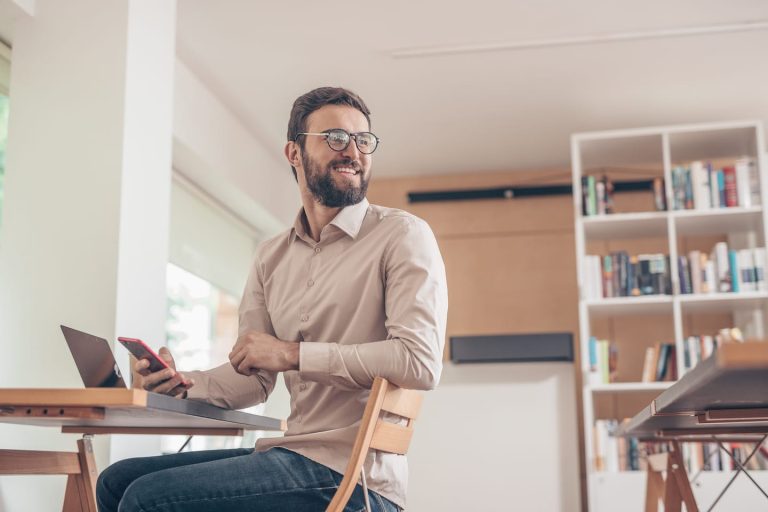A man with glasses and a beard is sitting in a modern office space, holding a phone and smiling. An open laptop is on the desk in front of him. Shelves filled with books are visible in the background.