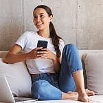 A woman sitting on a couch, smiling and holding a smartphone. She is casually dressed in a white t-shirt and blue jeans, with a laptop beside her. The background features a light-colored wall.