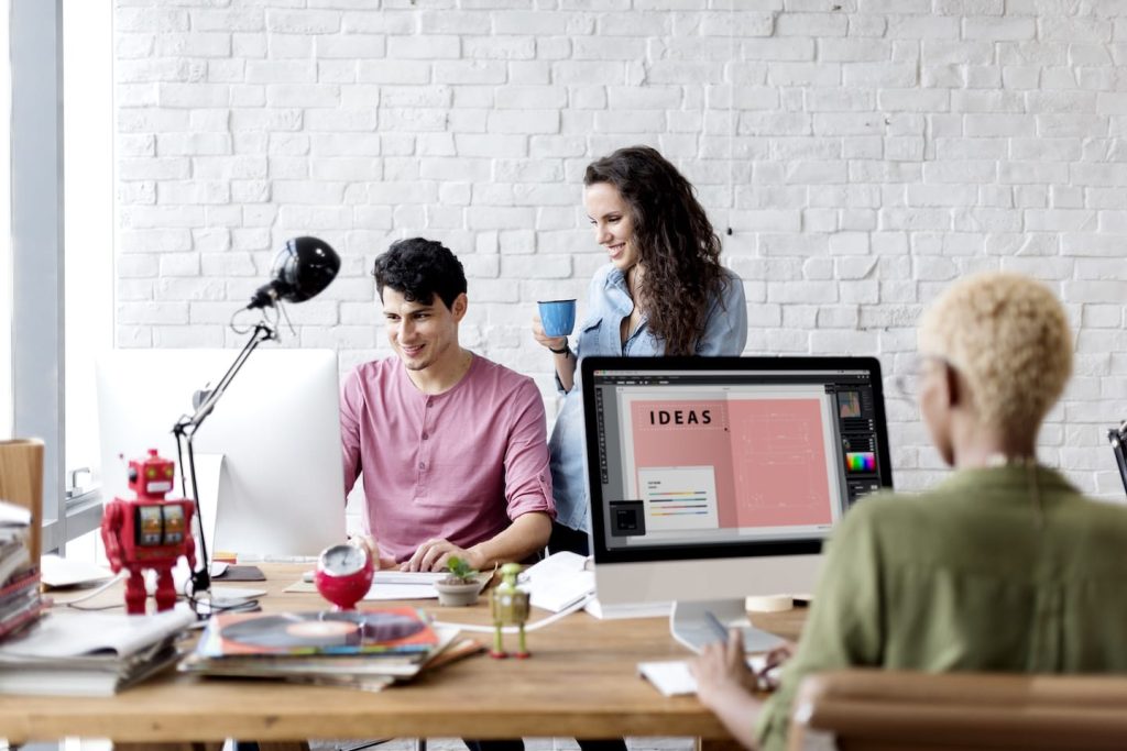 Three people in a modern office space. Two are discussing work at a desk with a lamp and robot figure, smiling at a computer. The third person, seen from behind, is working on a large monitor displaying the word IDEAS.