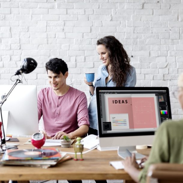 Three people in a modern office space. Two are discussing work at a desk with a lamp and robot figure, smiling at a computer. The third person, seen from behind, is working on a large monitor displaying the word IDEAS.