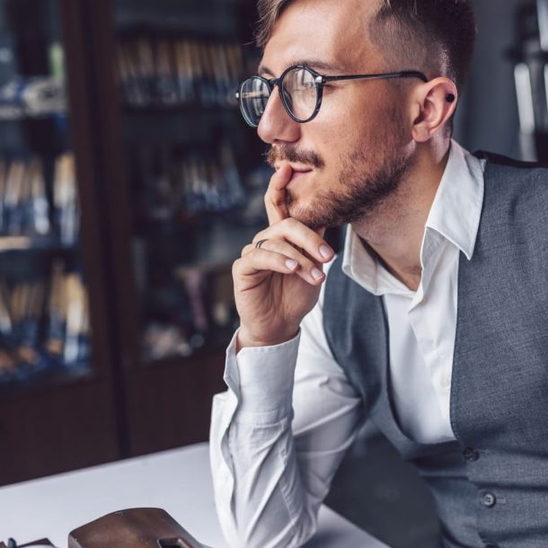 A man with glasses and a beard sits pensively at a table, contemplating his goals. Hes wearing a white shirt and grey vest, with a vintage typewriter and an open book in front of him. Bookshelves are in the background.