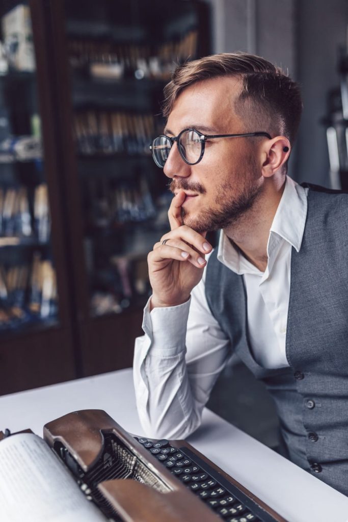 A man with glasses and a beard sits pensively at a table, contemplating his goals. Hes wearing a white shirt and grey vest, with a vintage typewriter and an open book in front of him. Bookshelves are in the background.