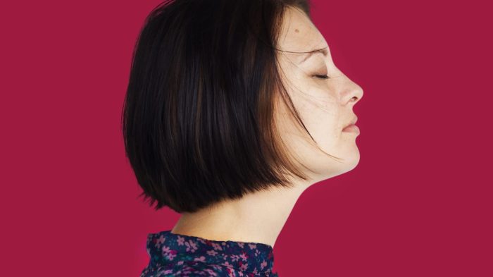 A woman with short dark hair is seen in profile against a solid red background. She has her eyes closed and appears to be serene. She is wearing a dark floral-patterned top.