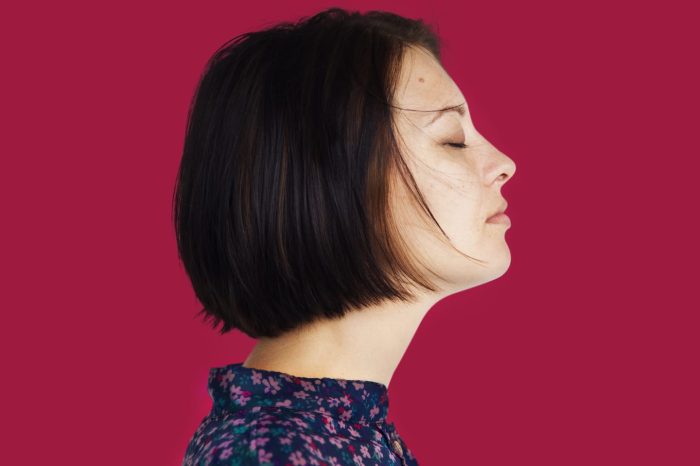 A woman with short dark hair is seen in profile against a solid red background. She has her eyes closed and appears to be serene. She is wearing a dark floral-patterned top.