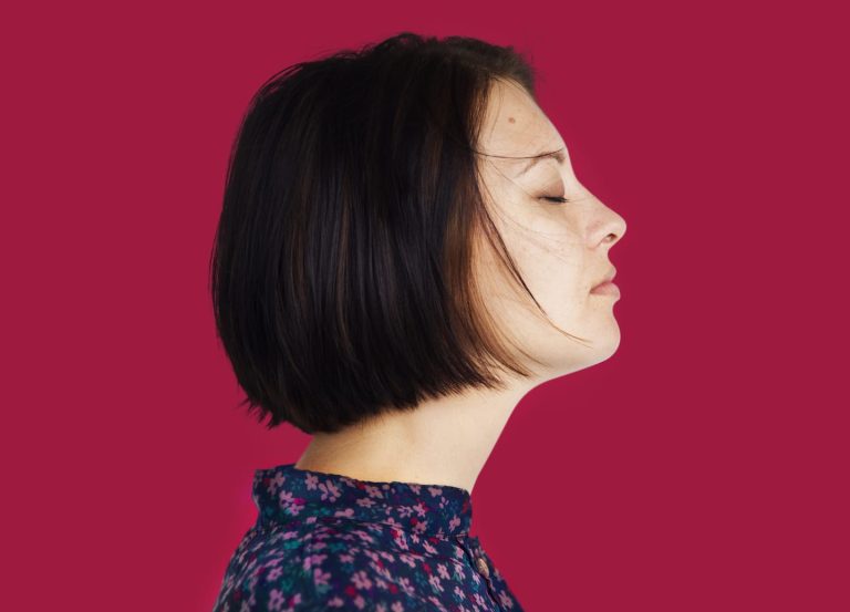 A woman with short dark hair is seen in profile against a solid red background. She has her eyes closed and appears to be serene. She is wearing a dark floral-patterned top.