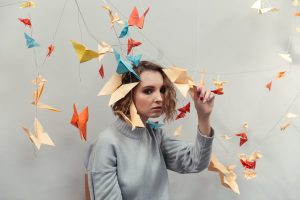 A person with wavy hair wearing a gray sweater sits surrounded by colorful origami birds hanging from strings. The background is a plain wall, and the individual is gently touching one of the origami pieces.