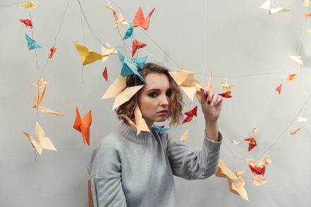 A person with wavy hair wearing a gray sweater sits surrounded by colorful origami birds hanging from strings. The background is a plain wall, and the individual is gently touching one of the origami pieces.