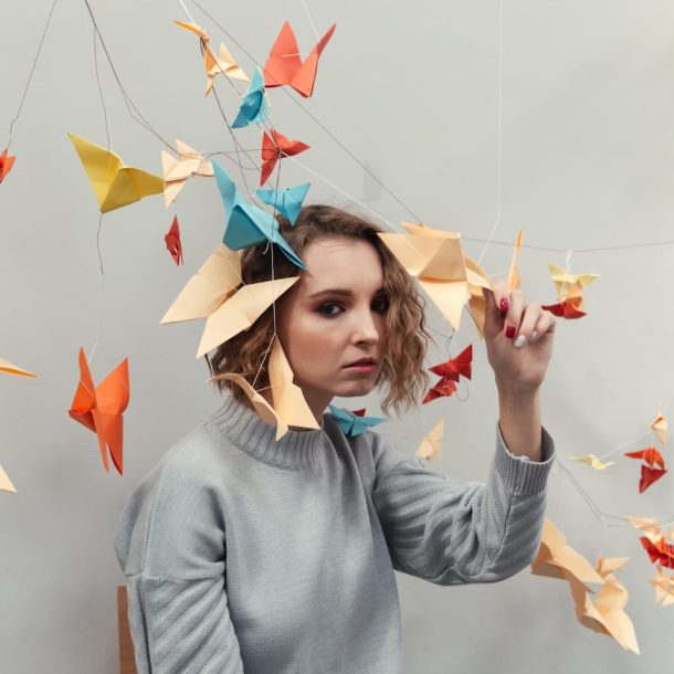 A person with wavy hair wearing a gray sweater sits surrounded by colorful origami birds hanging from strings. The background is a plain wall, and the individual is gently touching one of the origami pieces.