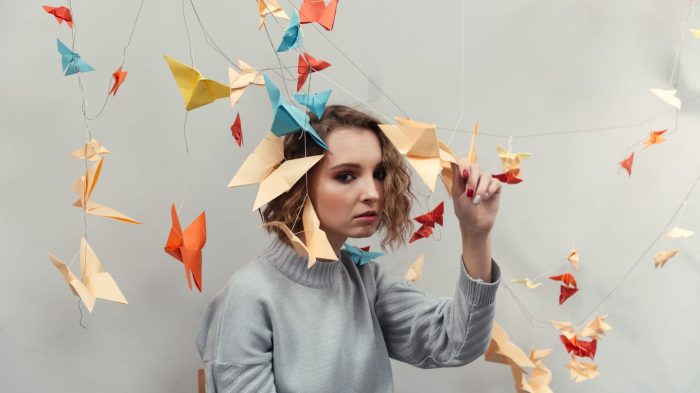 A person with wavy hair wearing a gray sweater sits surrounded by colorful origami birds hanging from strings. The background is a plain wall, and the individual is gently touching one of the origami pieces.