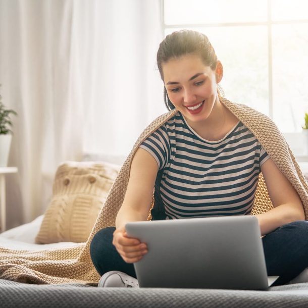 A woman sits cross-legged on a bed with a knitted blanket draped over her shoulders, smiling as she looks at a laptop. The room is bright, with a plant and a lamp on the bedside table. Natural light streams through the window behind her.